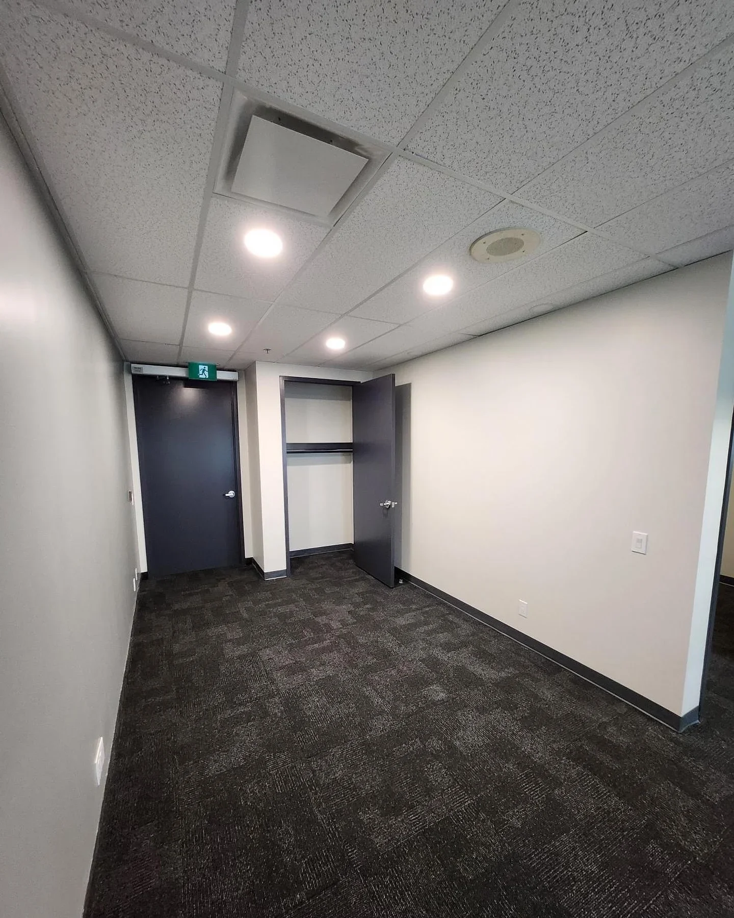 Empty office hallway with carpeted floors, white walls, closed black door with an exit sign, open closet, and ceiling with recessed lighting and vents.