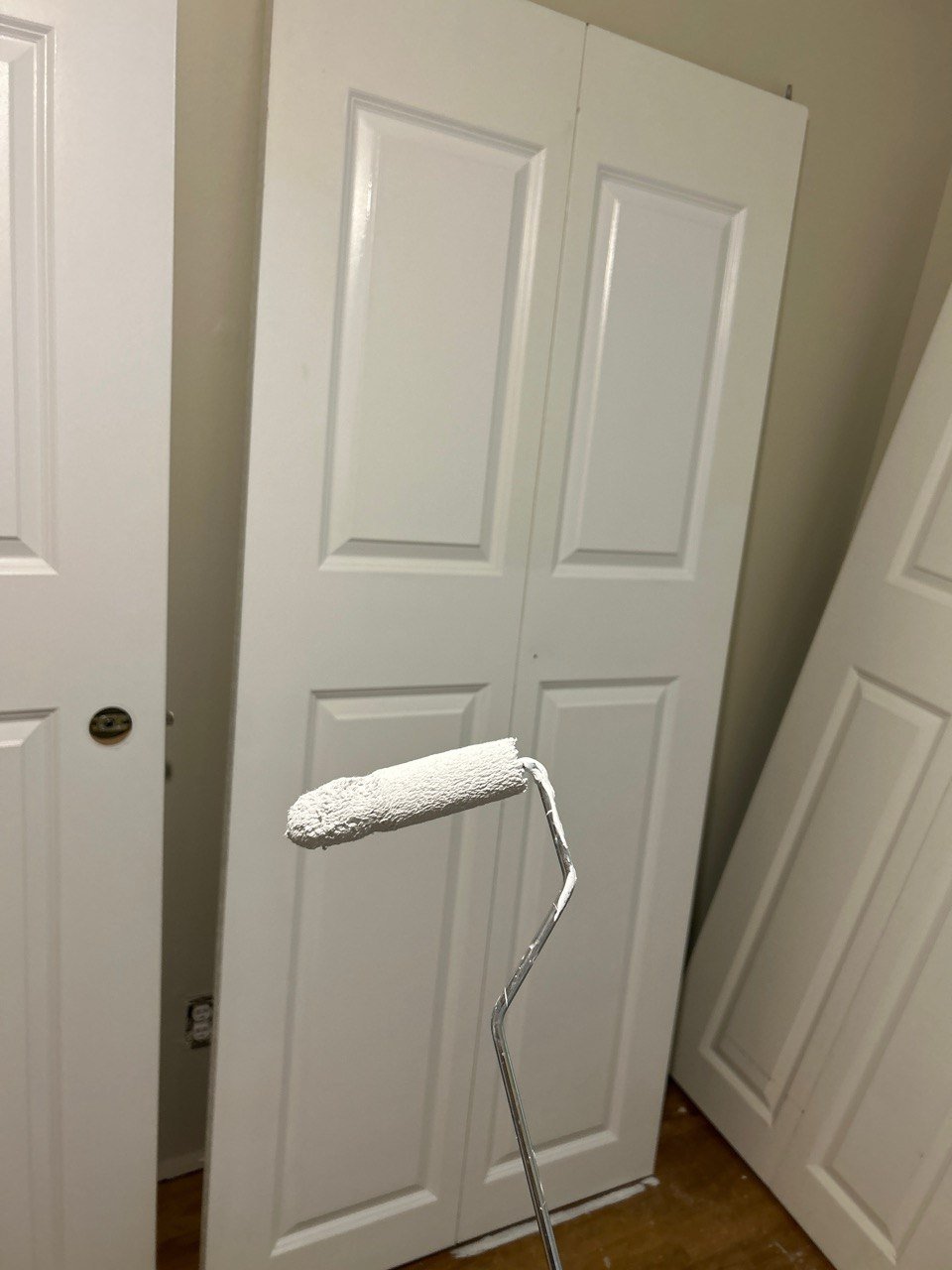 A white paint roller covered in white paint, held in front of a white closet door in a room with light-colored walls.