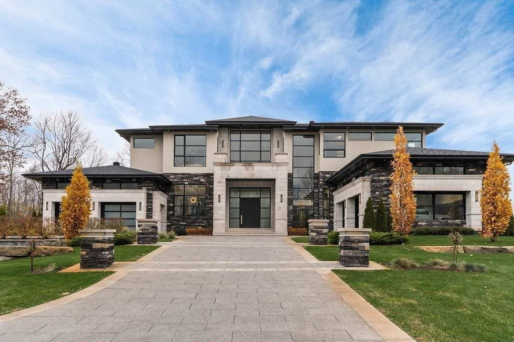 Modern large house with a stone and stucco exterior, a wide driveway, and landscaped front yard with trees and grass.