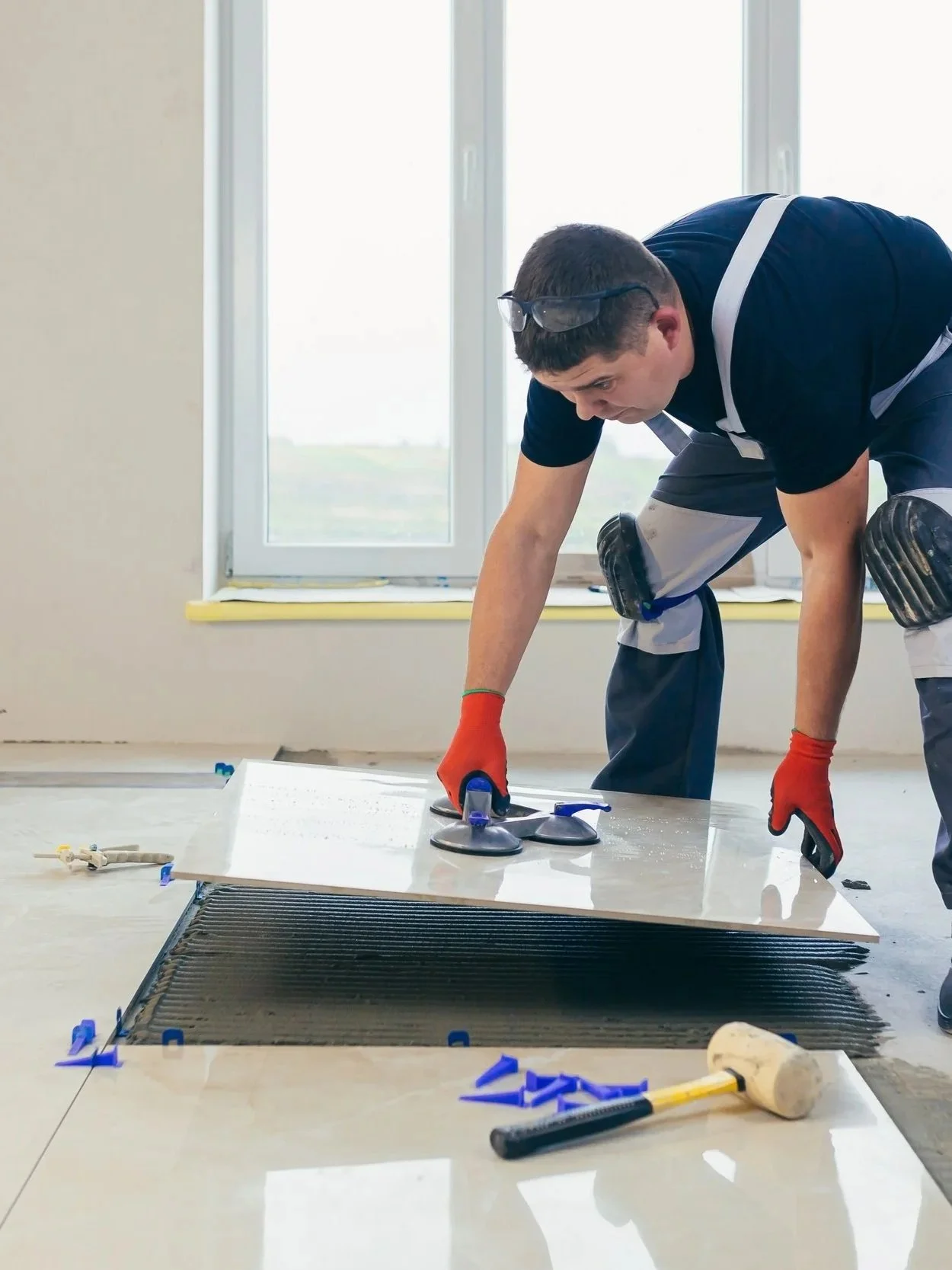 A man installing a large vent or HVAC grille on a floor, kneeling with tools nearby, wearing gloves and safety glasses on his head, in a room with large window and plain walls.