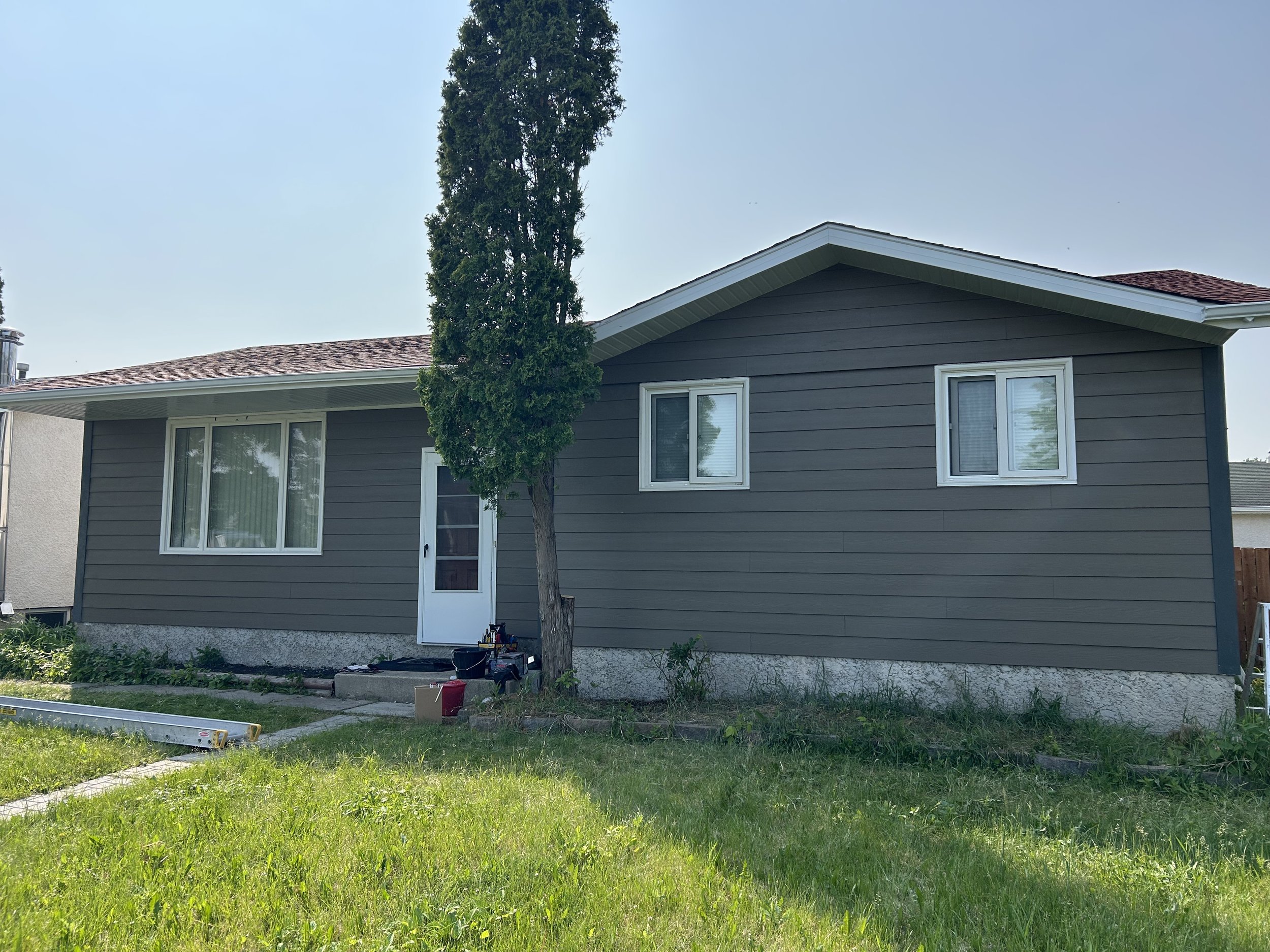A two-story house with gray siding, white window frames, and a brown shingled roof, with a tall, slim tree in front. There are tools and construction materials on the ground and steps leading to a screened door.