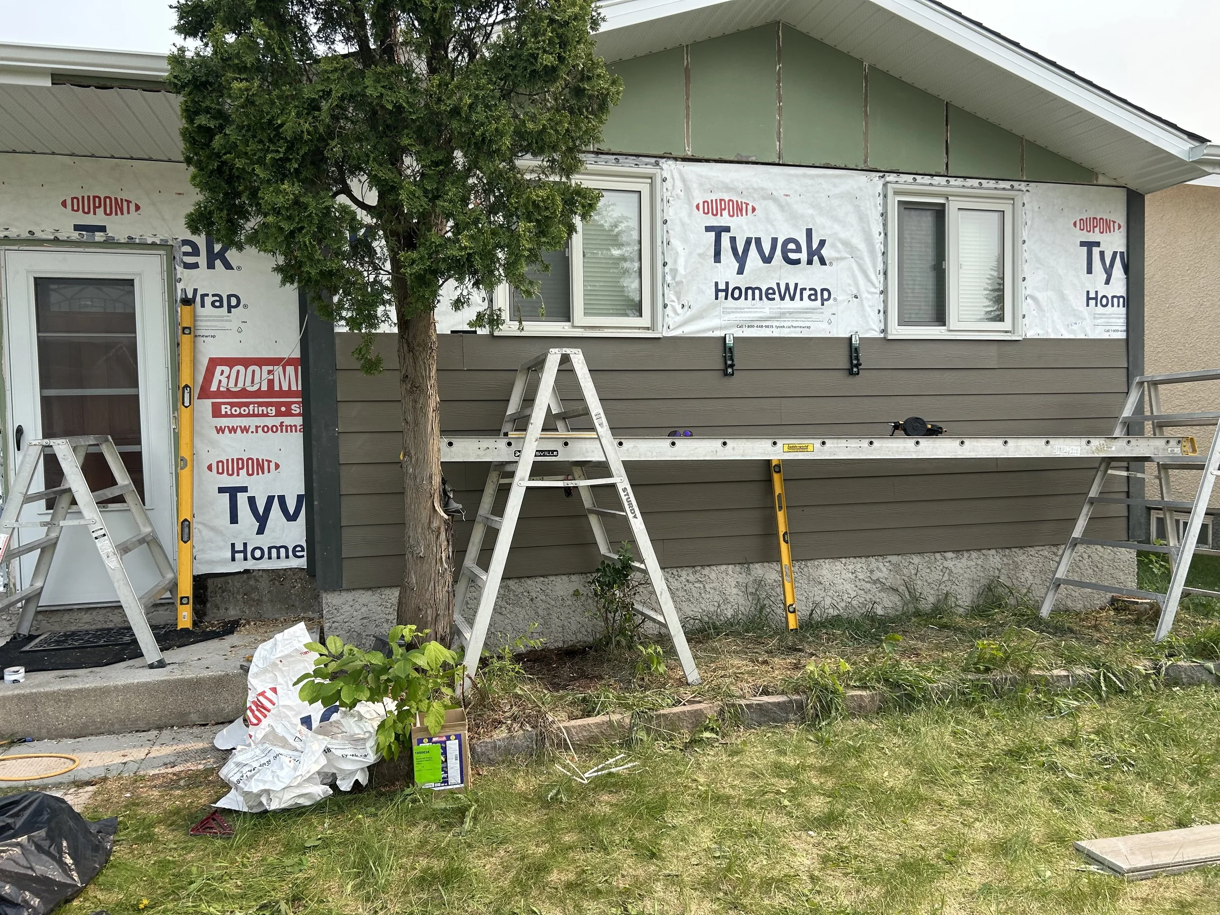 Construction site of a house exterior with partially installed siding, windows, and roofing materials. There are multiple ladders, a tree in front, and tools and materials scattered on the ground.