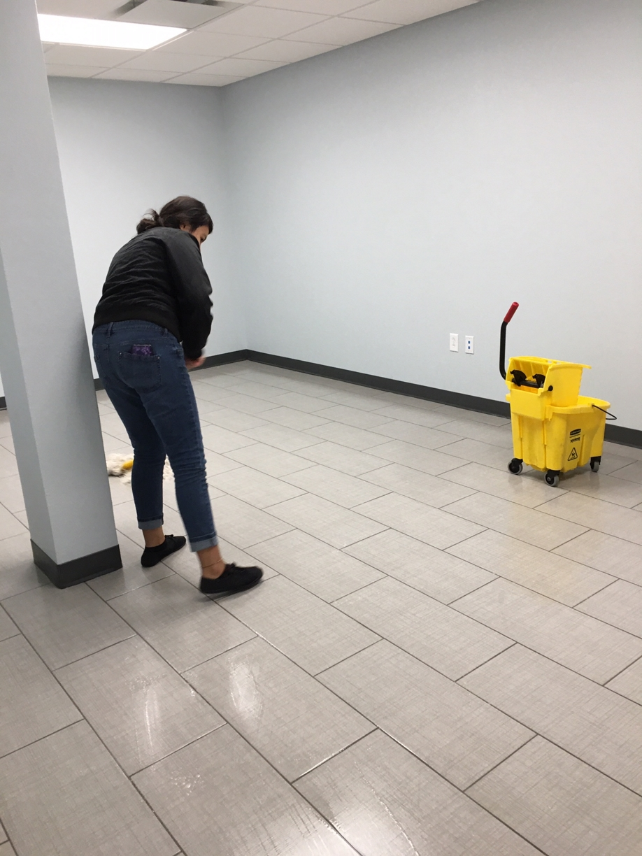 A woman mopping a tiled floor in an empty room with light blue walls and a yellow cleaning cart.
