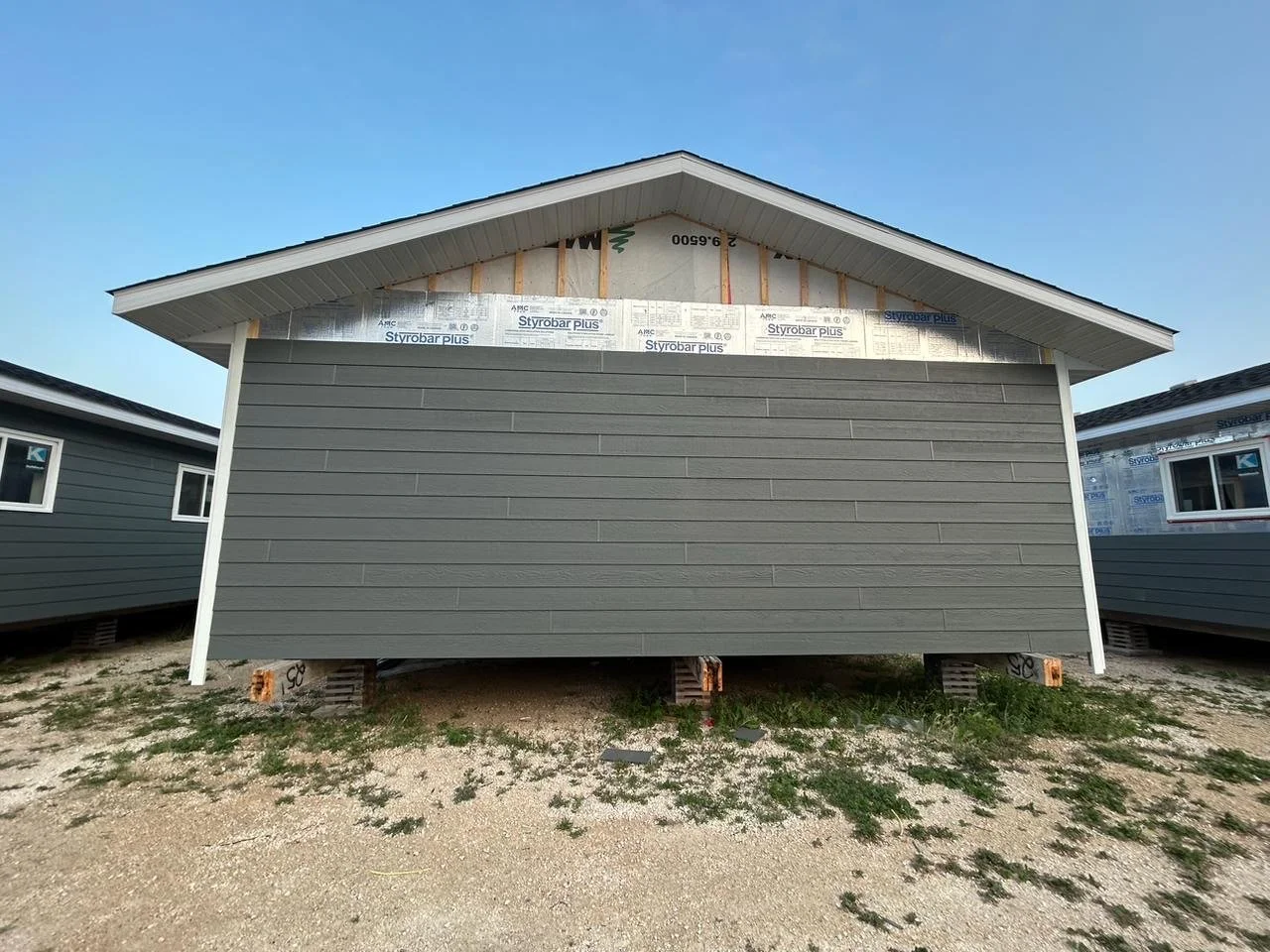 A house under construction with gray siding, elevated on concrete blocks, with the roof partially finished and some construction materials visible underneath, against a clear blue sky.