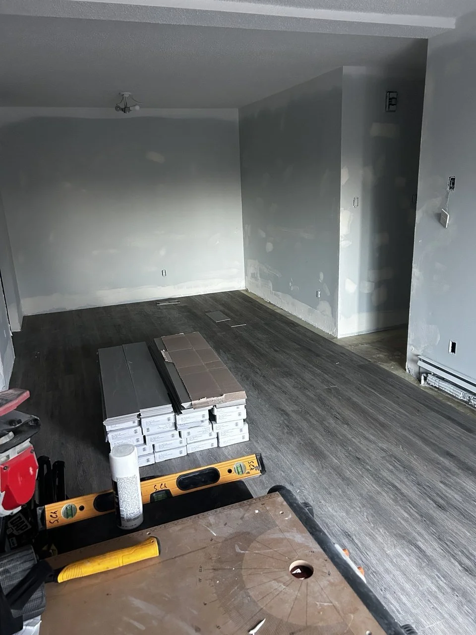 Room under renovation with partially installed dark wooden flooring, gray unfinished walls, and construction tools and supplies on the floor.