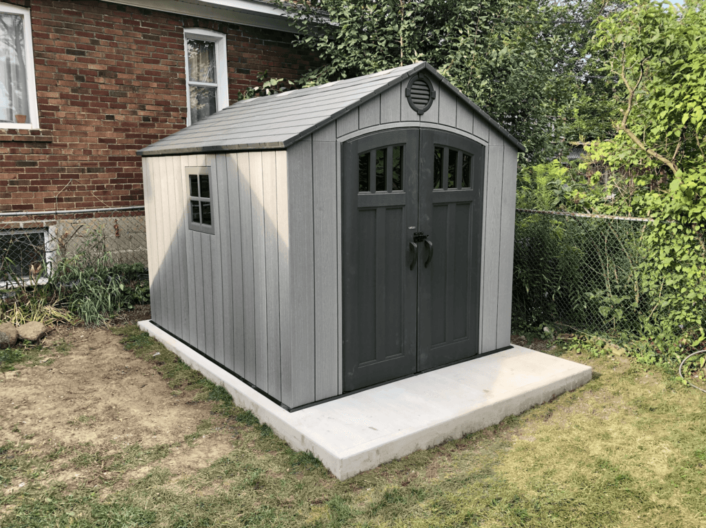 A gray outdoor shed with a double door, small windows, and a gable roof, situated on a concrete slab in a backyard with a brick house, a chain-link fence, and greenery.