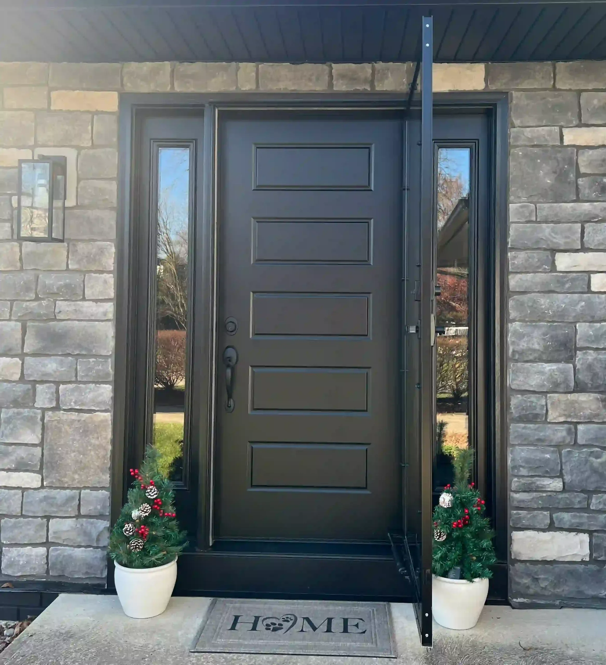 Black front door with decorative panels, flanked by two potted Christmas plants with red berries and pine cones, on a concrete porch with a welcome mat that says 'HOME' and has paw prints. The door has a black metal storm door in front of it, and there's a black post supporting a small black roof or awning above. The house has a stone brick exterior, and reflections of trees and sky are visible in the glass panes on the door.