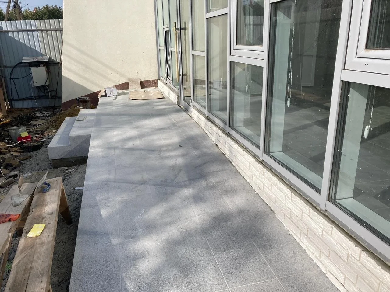 Construction site with a newly installed gray tiled patio beside a house with large glass sliding doors. Tools and construction materials are visible on the side, indicating ongoing work.