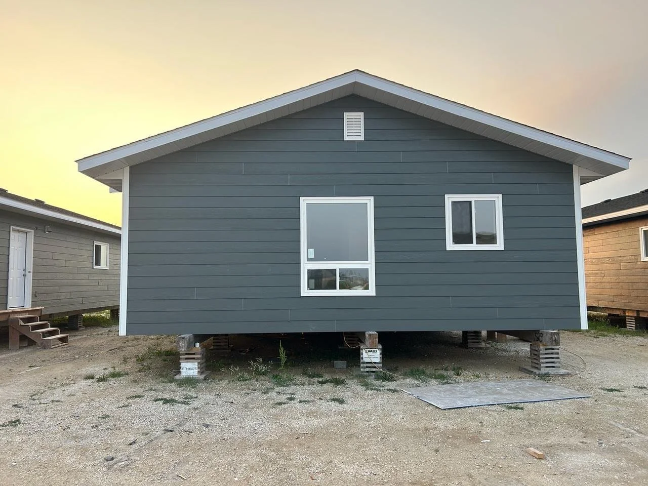 Unfinished blue house on stilts with white framed windows, located on a construction site with dirt ground and other similar houses in the background during sunset.