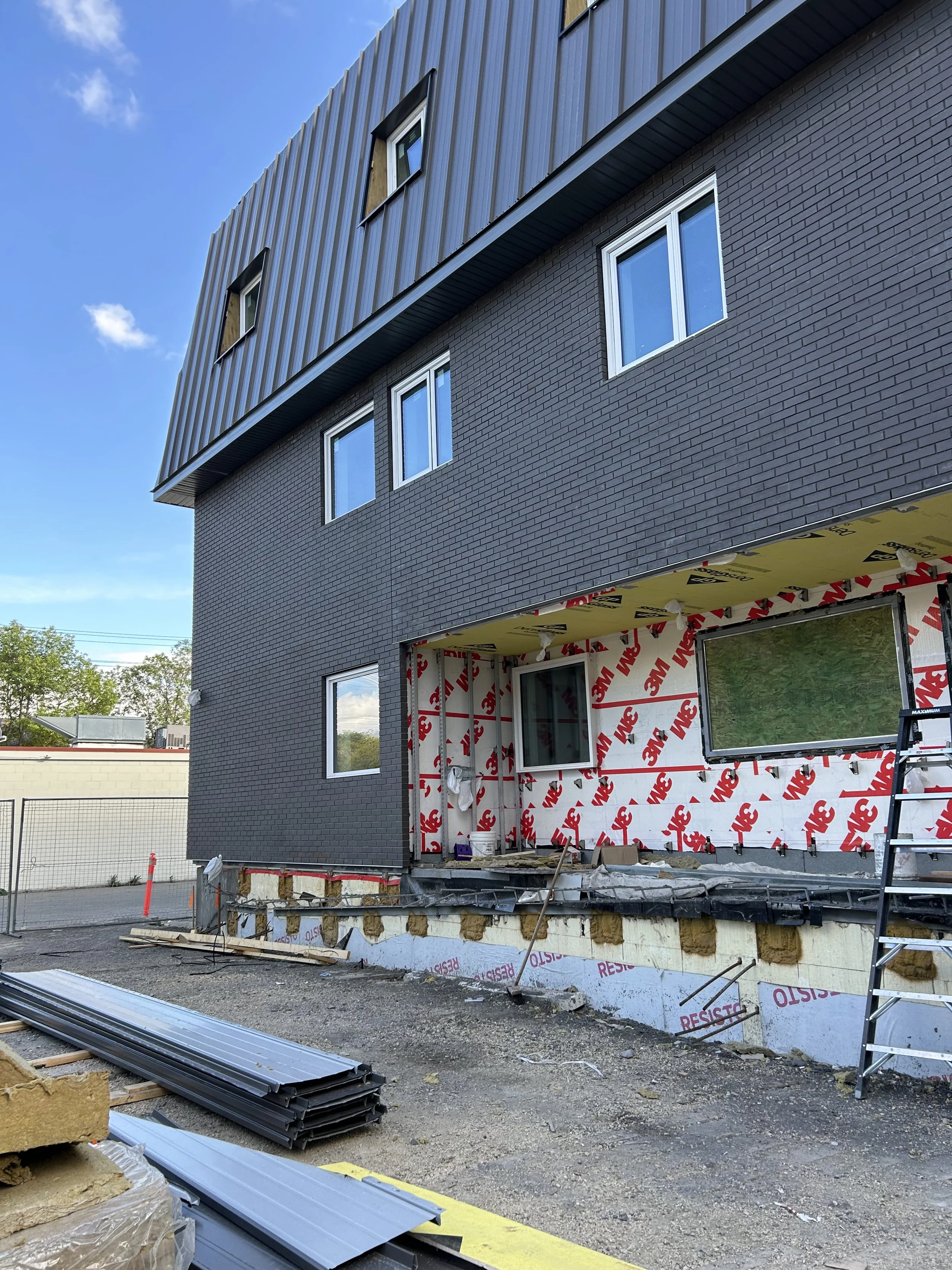 A building under construction with installed dark gray brick siding and several windows. The lower part of the building shows construction materials and insulation, with some areas still unfinished. A ladder is present on the right side.