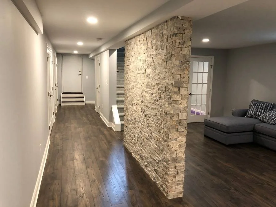 A modern basement interior with wood flooring, a stone accent wall, a gray sectional sofa, and a door leading outside or to another room.