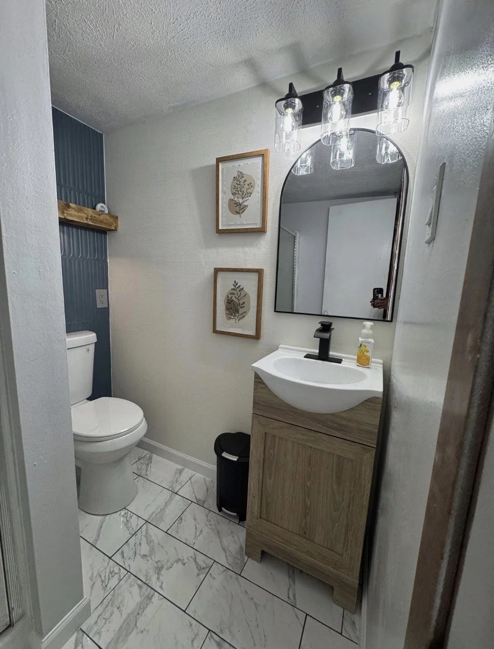 Small bathroom with white walls and white marble floor tiles. Features a toilet, a wood-finish vanity with a vessel sink, a black faucet, and a large mirror. Above the mirror, there are three glass light fixtures. Two framed botanical prints hang on the wall, and a wooden shelf with a small white item is on the wall near the toilet.