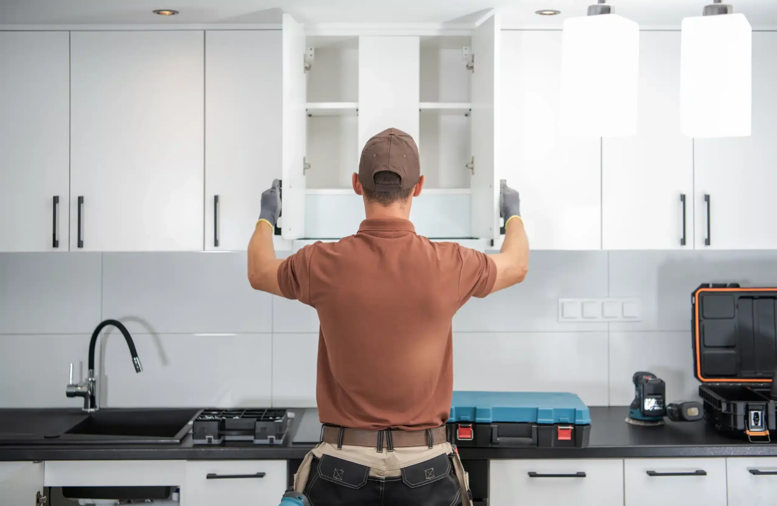 A man wearing a brown cap, brown shirt, black pants with white stitching, and gray gloves is opening a white kitchen cabinet.