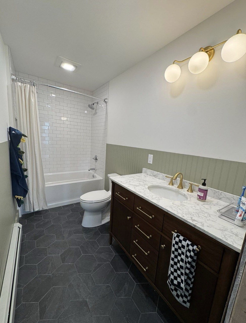 Bathroom with a bathtub and shower, toilet, dark wood vanity with marble countertop, gold fixtures, black and white checkered towel, white subway tile shower, green wainscoting, black hexagonal floor tiles, wall-mounted light fixture with four globe lights, bottle of lotion, and other toiletries.