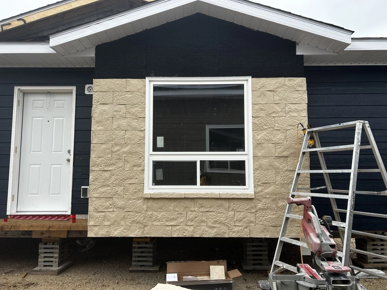 Exterior of a house under renovation, with a beige stone facade, a white door, and a large window. A ladder and construction tools are present, and the house is elevated on cinder blocks.