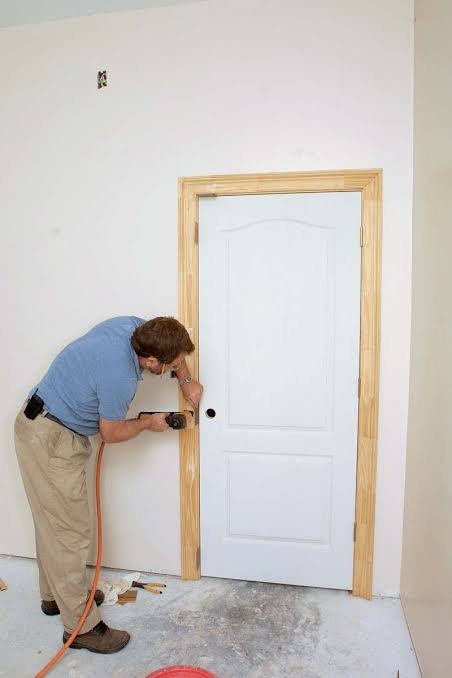 A man installing a white interior door with a natural wood frame in a room during construction or renovation.