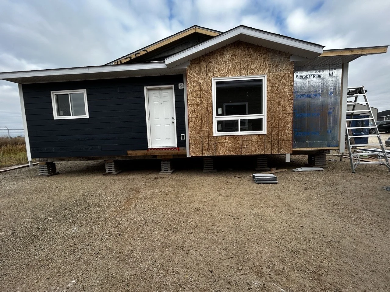 A house under construction with one side painted dark blue, and the other side with plywood and insulation visible. The house is elevated on concrete blocks, with a ladder and construction materials nearby.