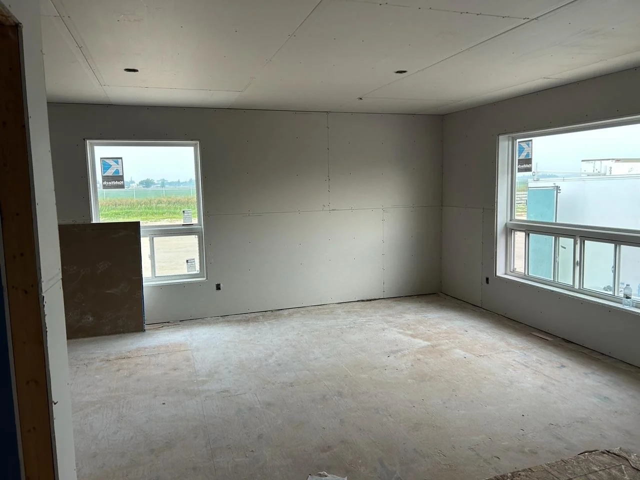 Empty room under construction with drywall on walls and ceiling, two large windows overlooking a grassy field and industrial area, unfinished flooring