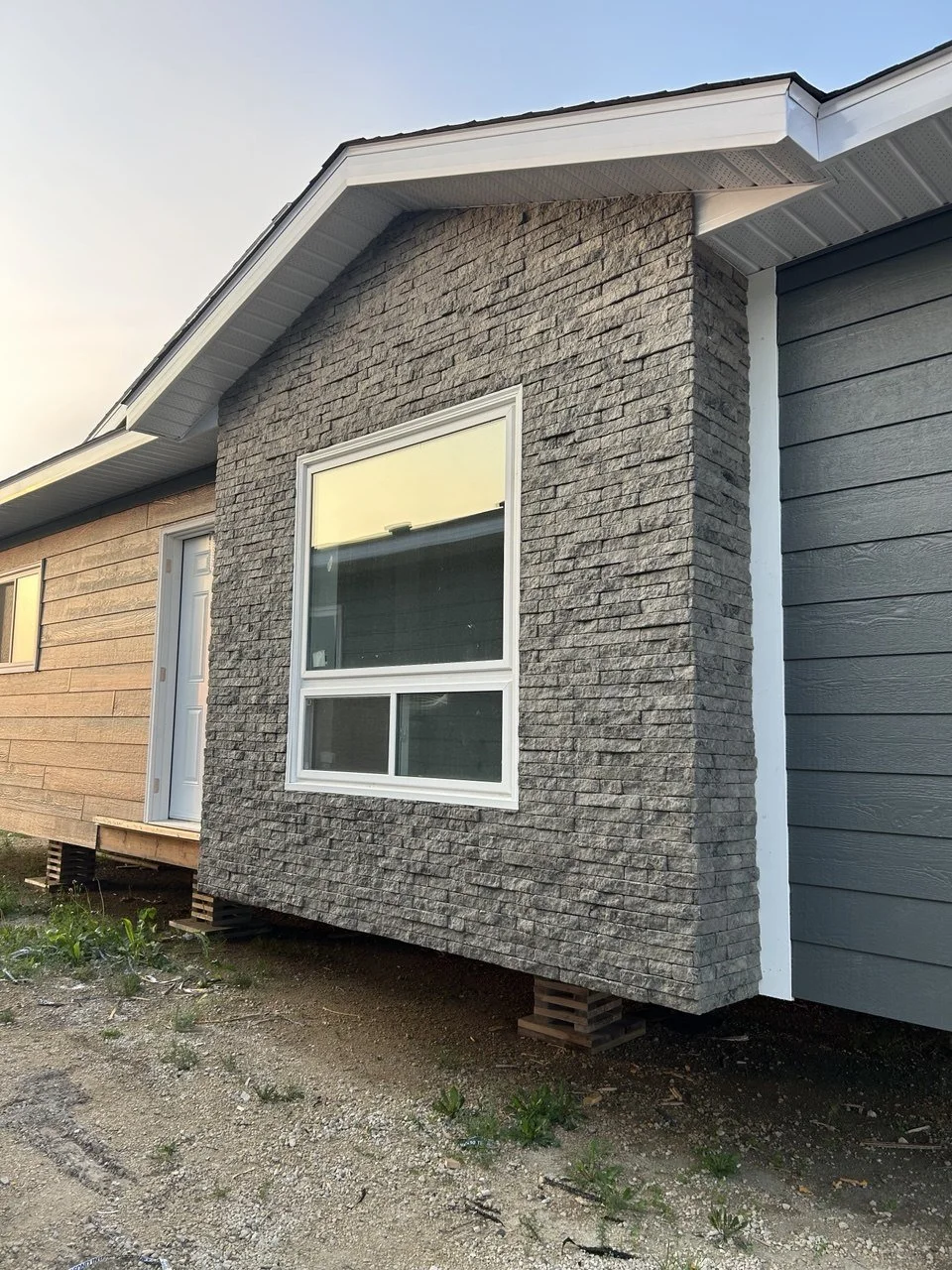 Close-up of a residential house exterior, showing a window, gray stone siding, and a section of gray painted siding, with the house elevated on wooden blocks.