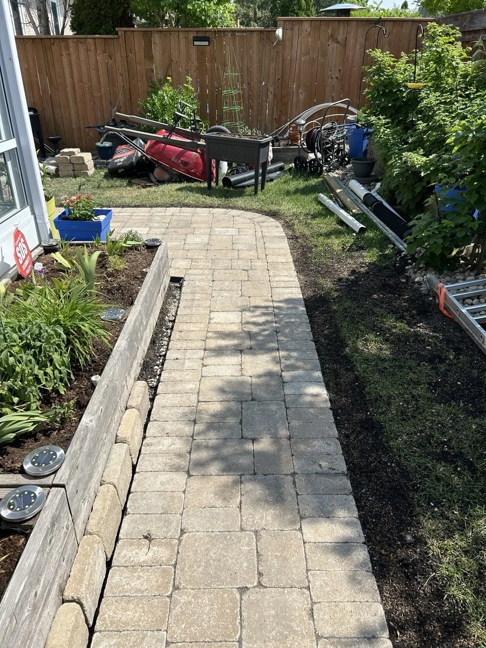 A backyard garden with a brick pathway, raised flower bed on the left with blooming plants, and various gardening tools and equipment on the right side near a wooden fence. Shadows of trees are cast on the pathway in bright sunlight.