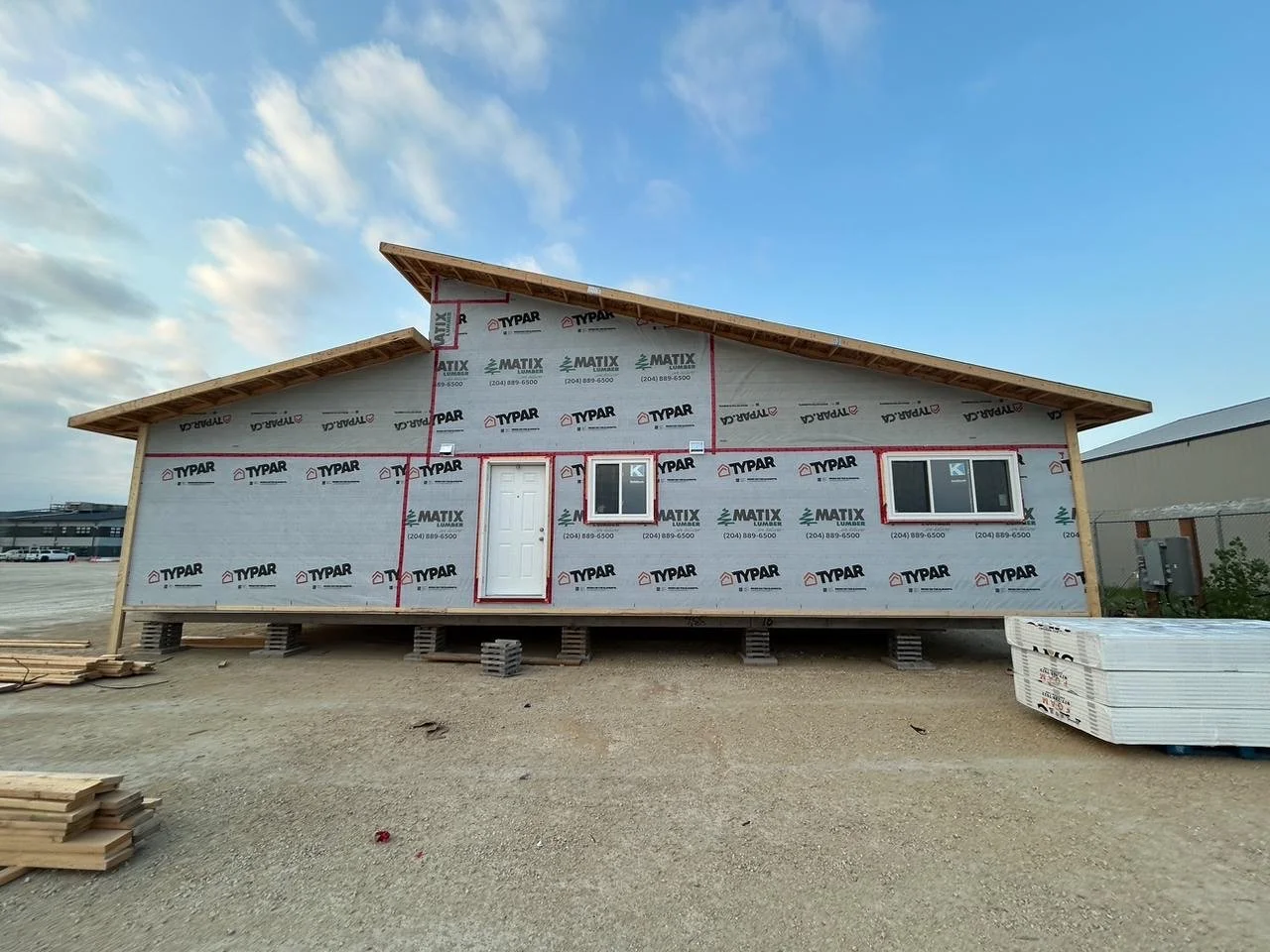 A house under construction with the exterior wrapped in Typar and Matix building wrap, elevated on concrete blocks, with a white door, two windows, and a sloped roof.