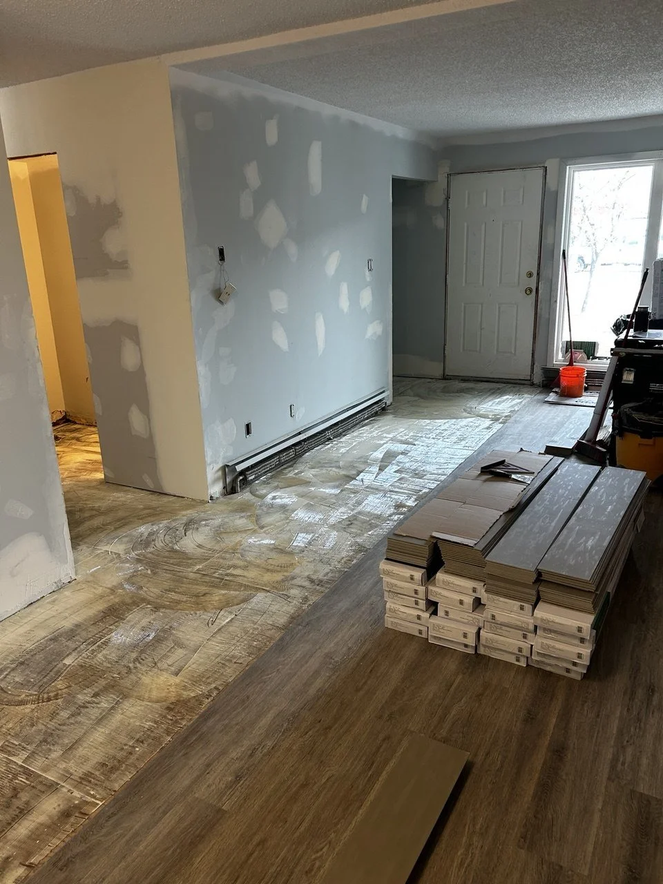 Interior of a room under renovation with partially installed flooring, new drywall patches on the walls, and stacks of flooring planks ready to be laid.