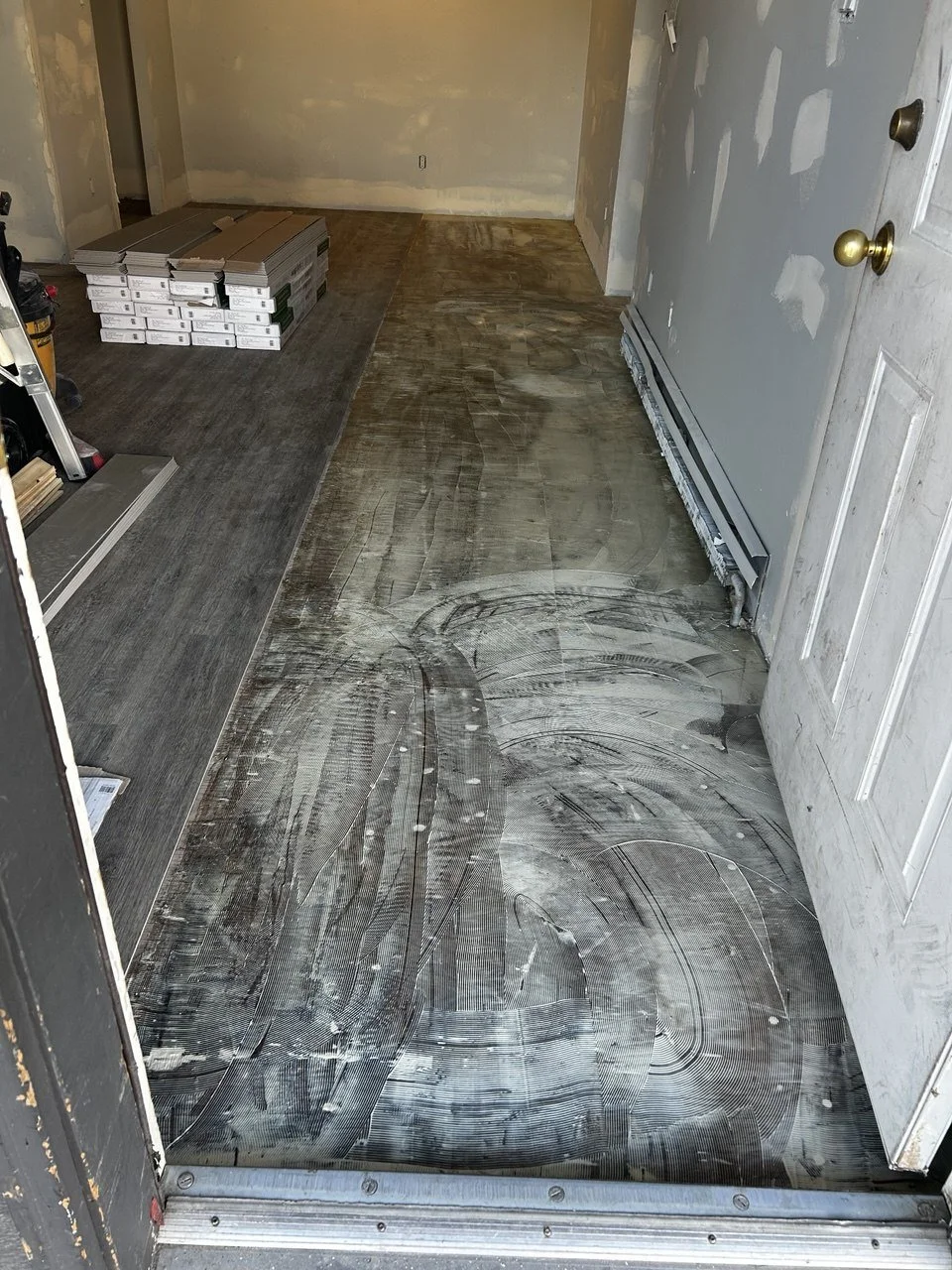 Inside a room under renovation with partially installed flooring. The left side has new flooring, while the right side has an unfinished subfloor covered in construction marks and debris. There are stacked boxes of tiles or flooring materials in the 