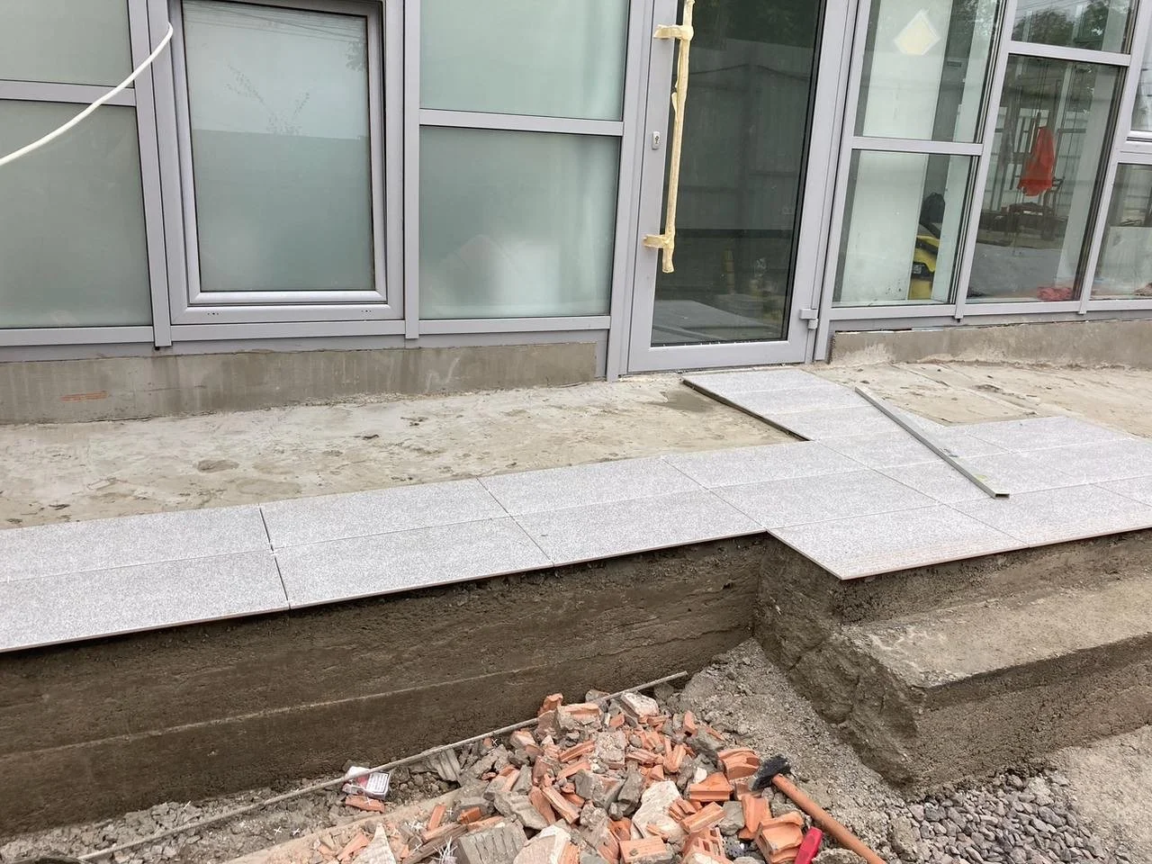 Construction site with new tiled walkway in front of glass building, dirt and rubble below, and step at the edge of the tiles.