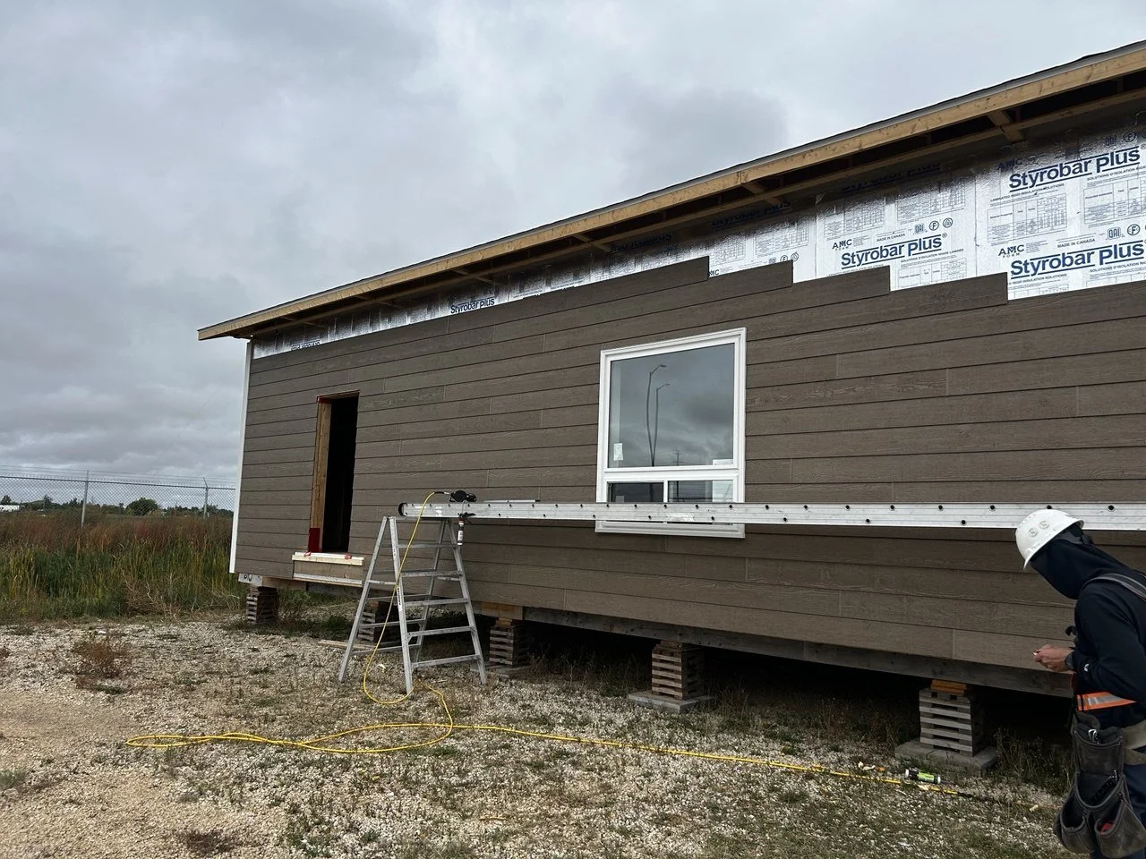 A house under construction with brown siding, a window, and an open door. Construction tools and materials are visible, including a ladder and a worker wearing a helmet and safety gear.