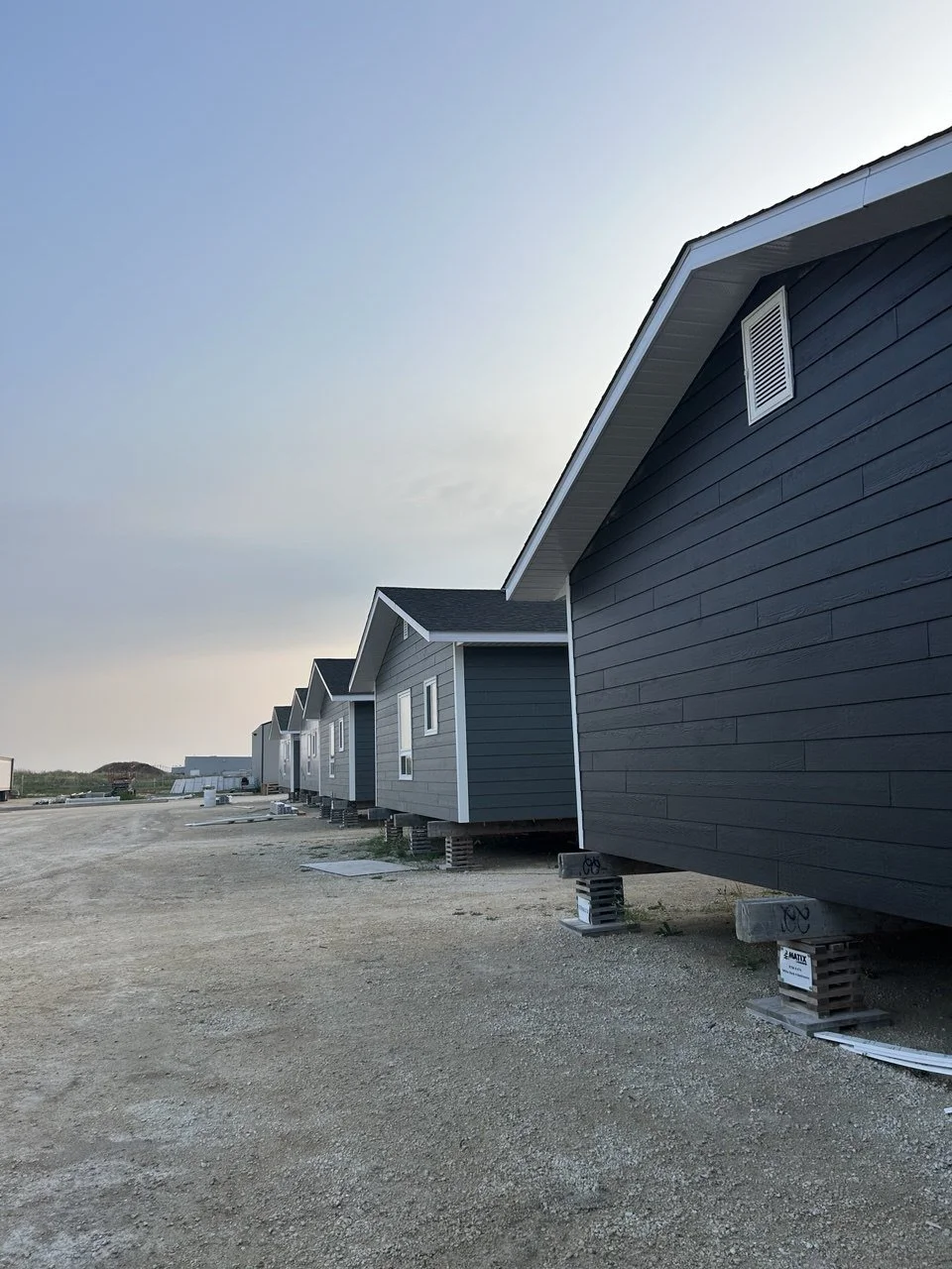Row of new gray houses under construction, elevated on concrete blocks, on a gravel lot.