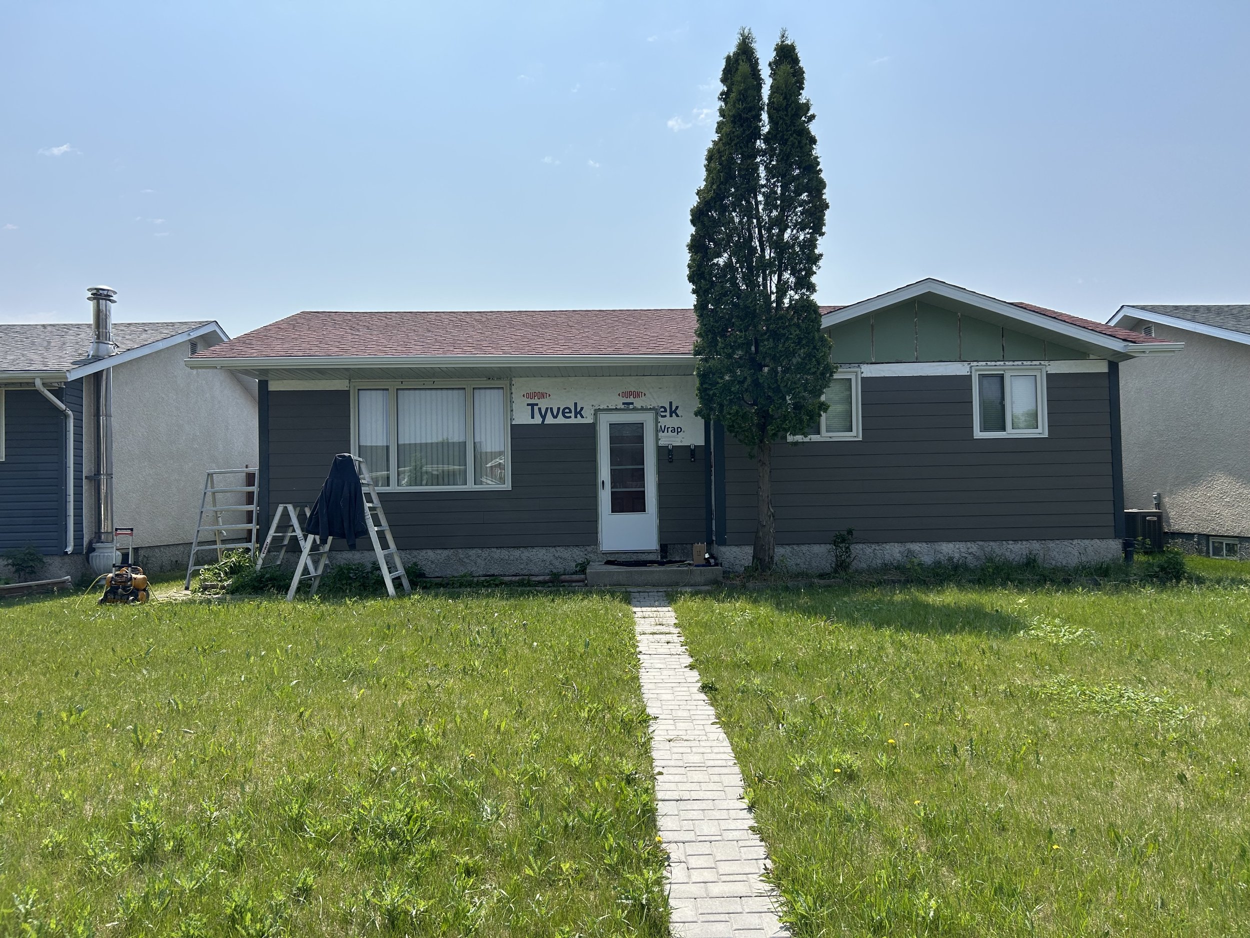 A backyard with a green lawn, a small brick pathway, and a multi-story house with gray and white siding, a red-tiled roof, window, and door. A tall, narrow tree stands in front of the house, and there are some construction materials and tools on the 