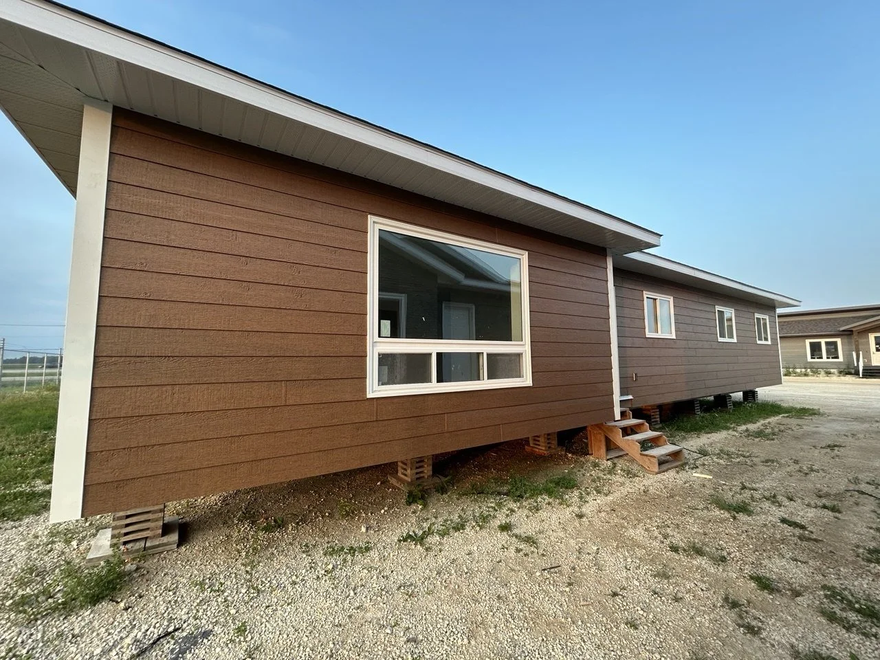 Exterior of a newly built raised house with brown and gray siding, small wooden stairs at the front, and gravel ground, under a clear blue sky.