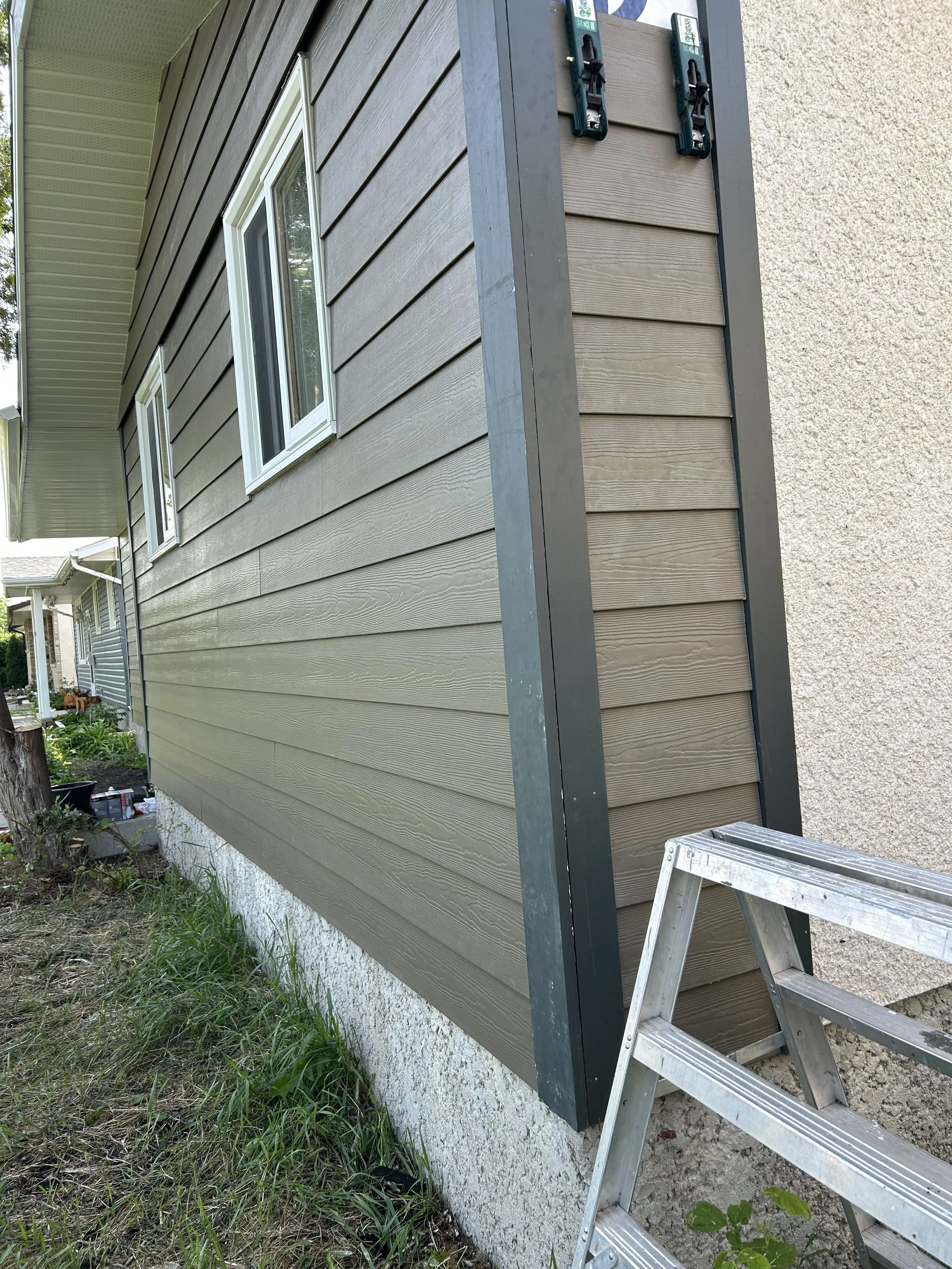 A house exterior wall with light brown horizontal siding, a window with white trim, an aluminum step ladder, and green construction clamps.