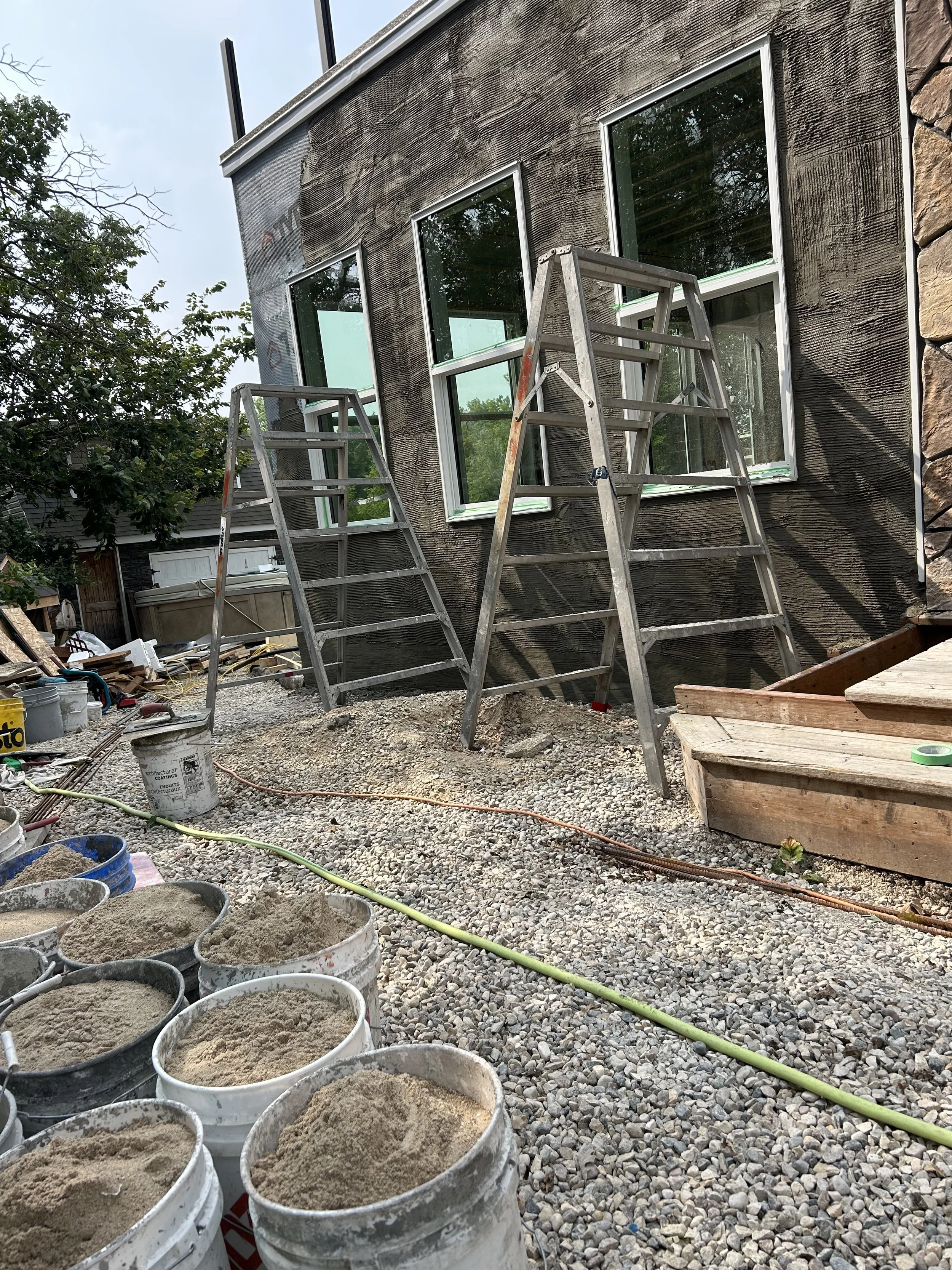 Under construction site showing buckets of sand in foreground, gravel ground, two ladders leaning against a building with three windows, partially covered with dark weatherproofing material, and construction tools and materials scattered around.