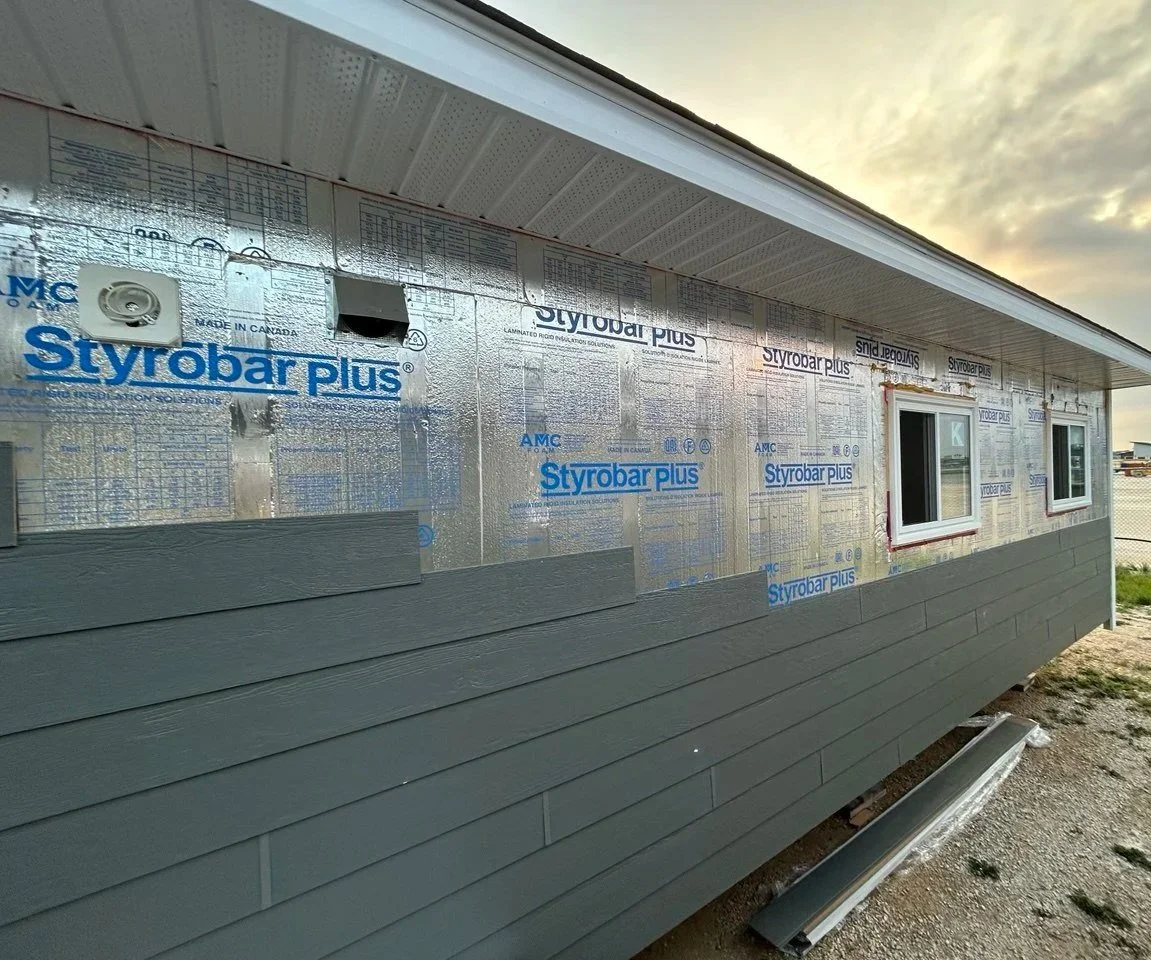 Side of a house under construction, with vinyl siding and insulation panels labeled 'Styrobar Plus' visible, along with two small windows and a portion of the roof and sky.