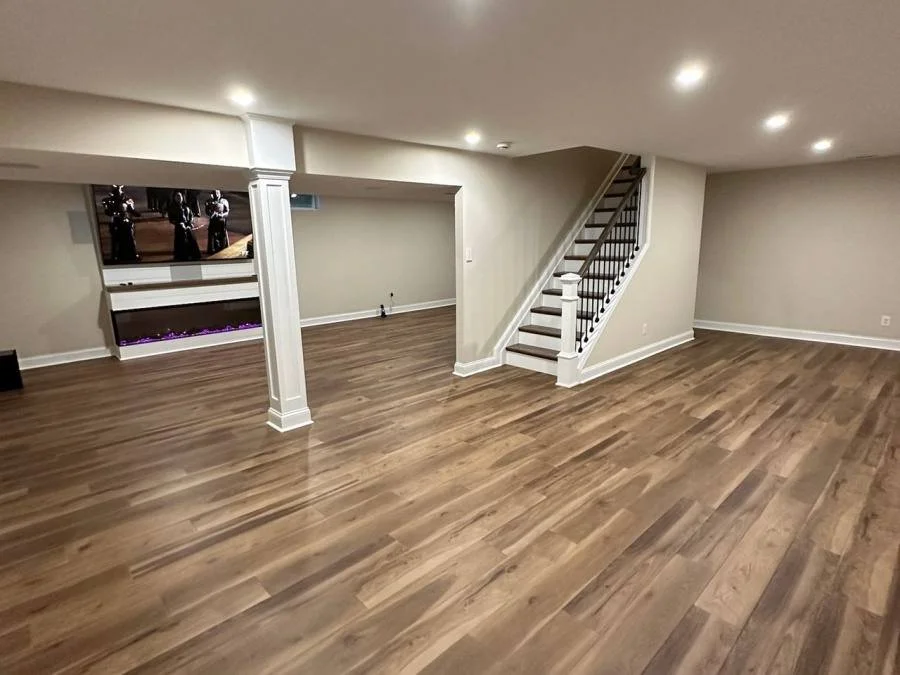 Empty finished basement with wood flooring, white walls, staircase, and recessed ceiling lights.