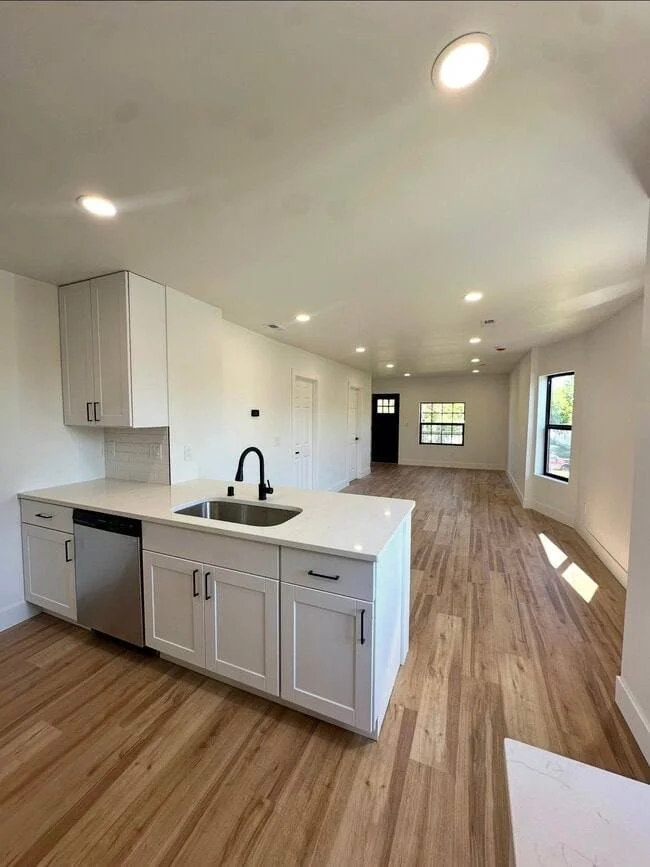 Empty open-concept kitchen and living area with white cabinets, a black faucet, hardwood floors, and large windows.