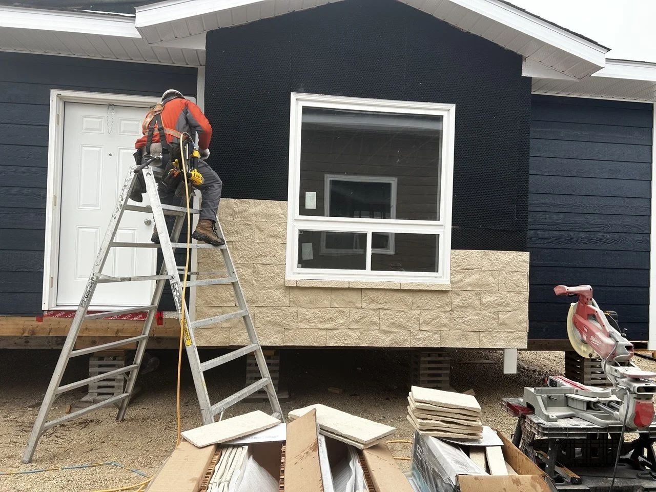 A construction worker on a ladder installing siding on a house with beige stone accents and a large window, with tools and construction materials around.