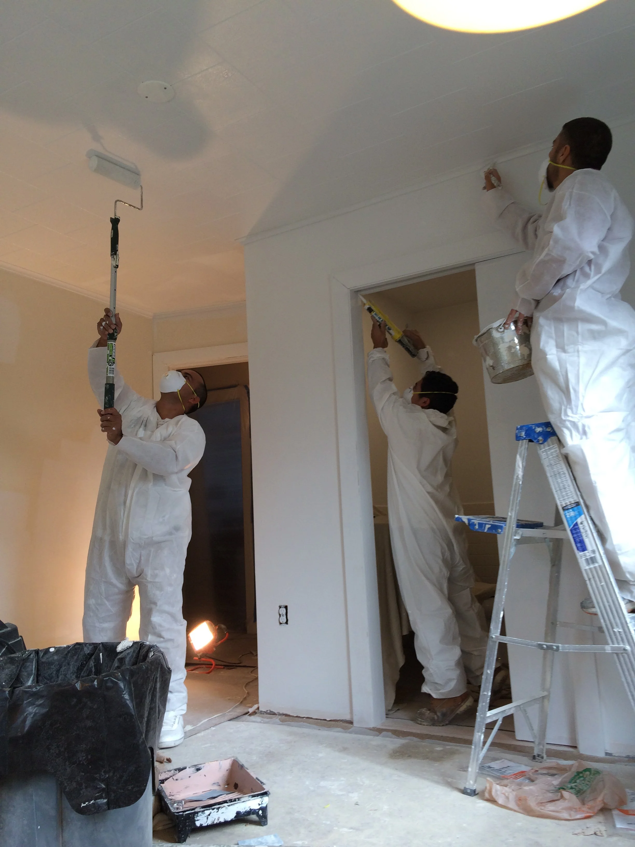 Four workers wearing white coveralls and masks painting a ceiling with paint rollers, while standing on a ladder and holding buckets, in a room undergoing renovation.
