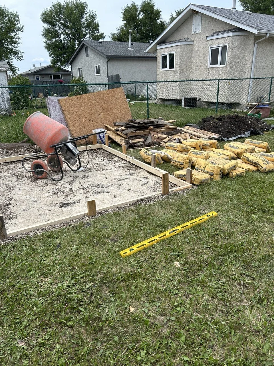 A backyard in the process of construction with a gravel base, a wheelbarrow, bags of insulation, wooden planks, and construction boards, enclosed by a chain-link fence with neighboring houses and trees in the background.