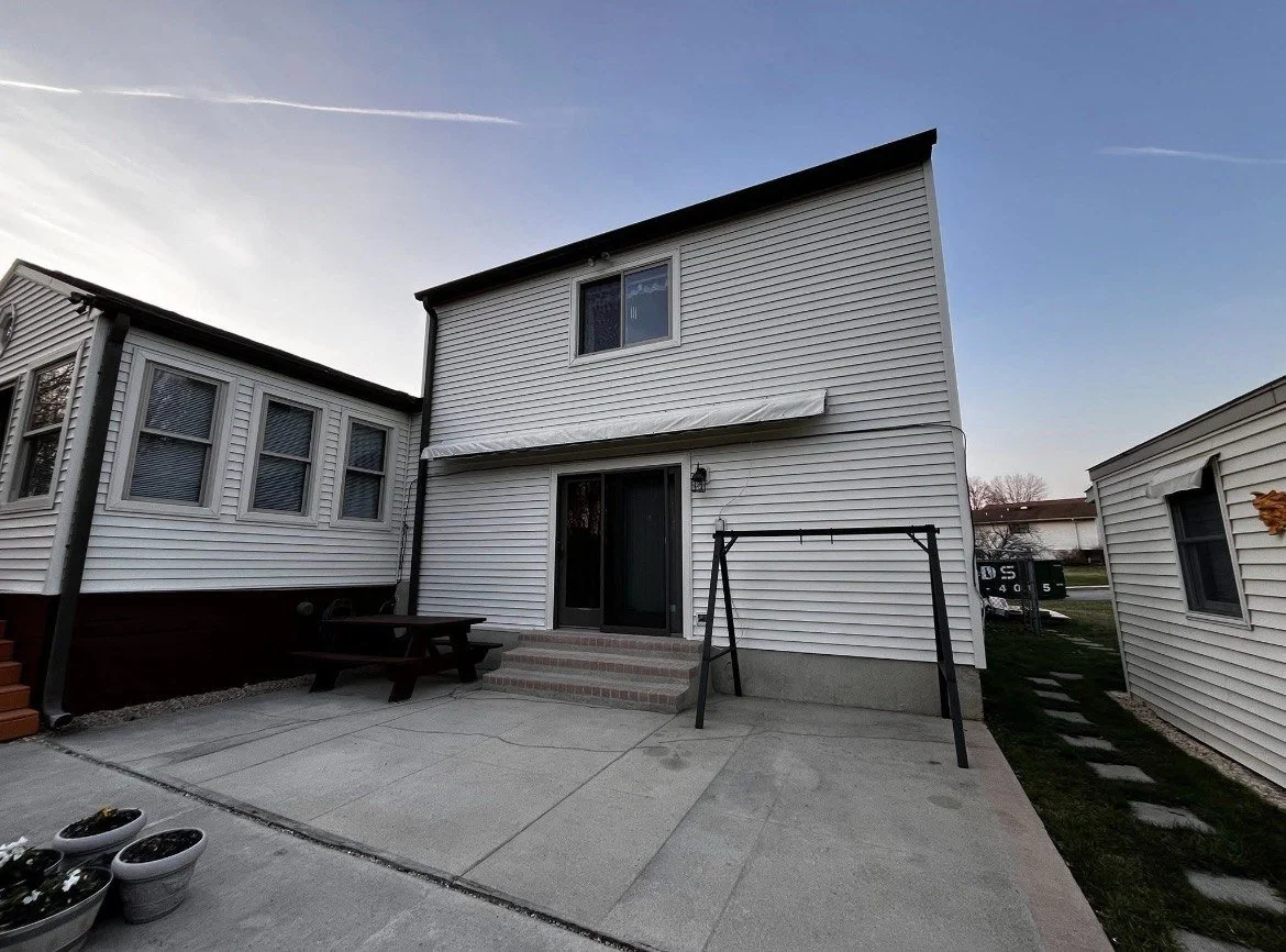 The back yard of a two-story white house with a sliding glass door, steps, and a black metal swing frame, with neighboring houses visible.