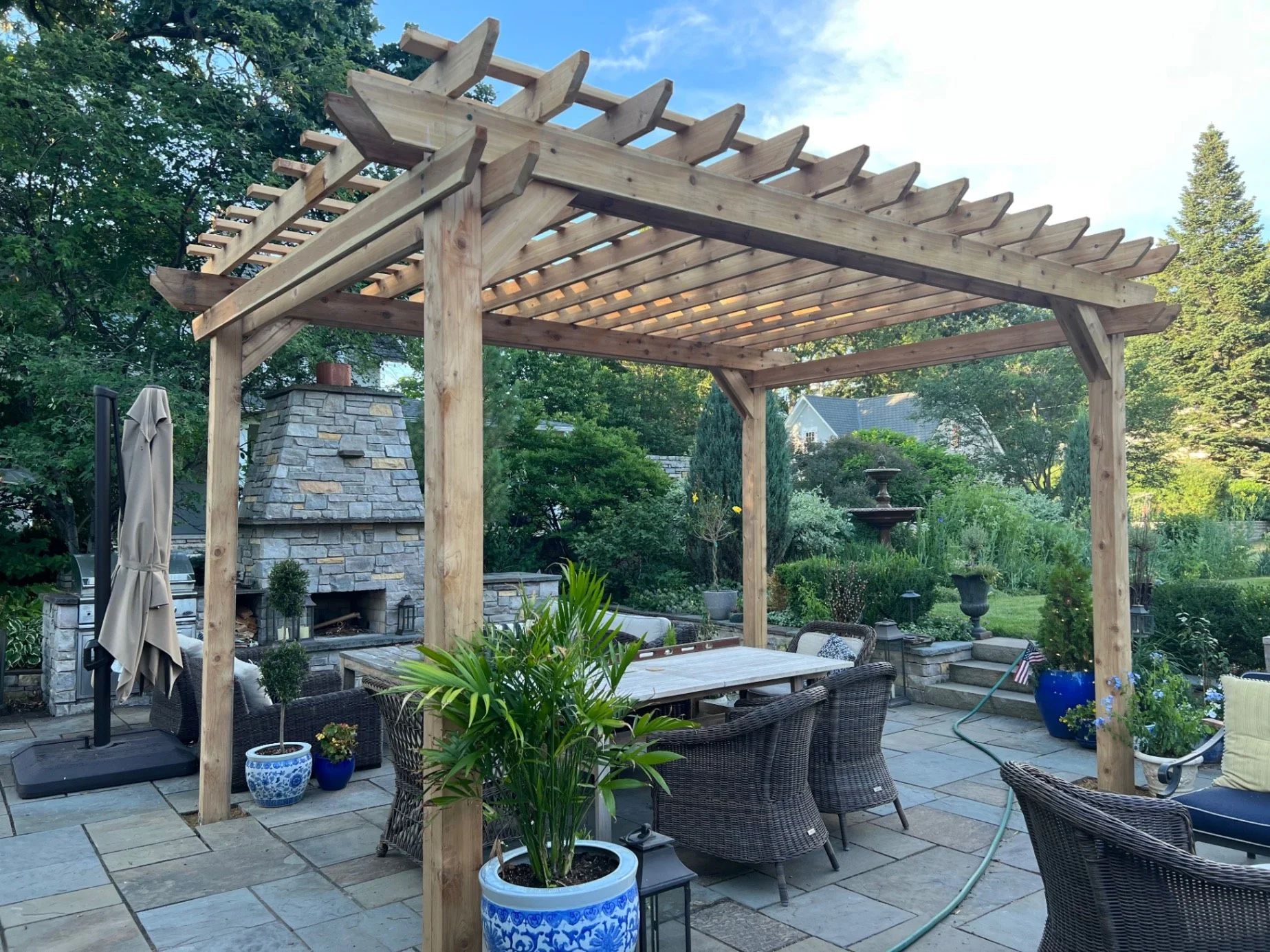 A backyard patio with a wooden pergola, outdoor dining table, wicker chairs, potted plants, a stone fireplace, and lush green trees in the background.