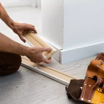 Person installing baseboard molding along a wall with a measuring tool.