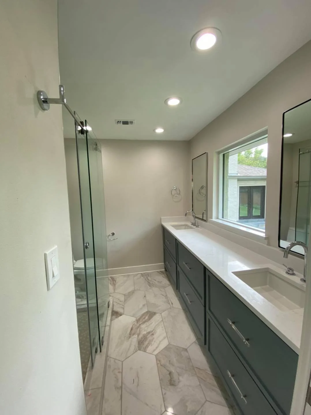 Modern bathroom with a large vanity, double sinks, granite countertop, large window, and a glass-enclosed shower with marble hexagonal tile flooring.
