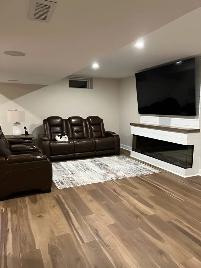 Living room with brown leather furniture, a flat-screen TV mounted above a white fireplace, a patterned rug, a small window, and a lamp on a side table.