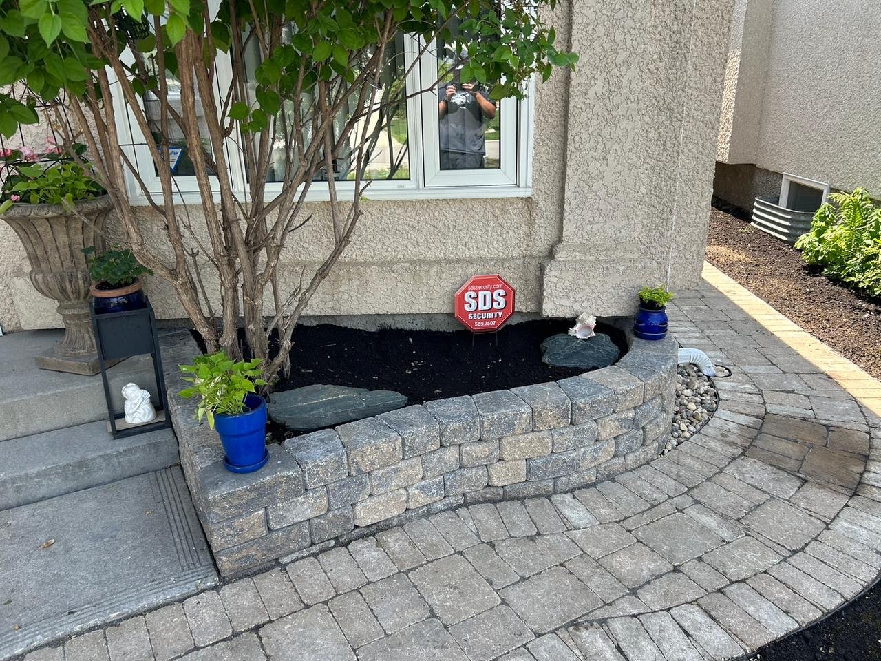 Front yard garden bed with a small tree, two blue flower pots with green plants, a black decorative lantern, a small white figurine, and a red security alarm sign, bordered by a curved stone brick wall surrounded by a brick walkway.