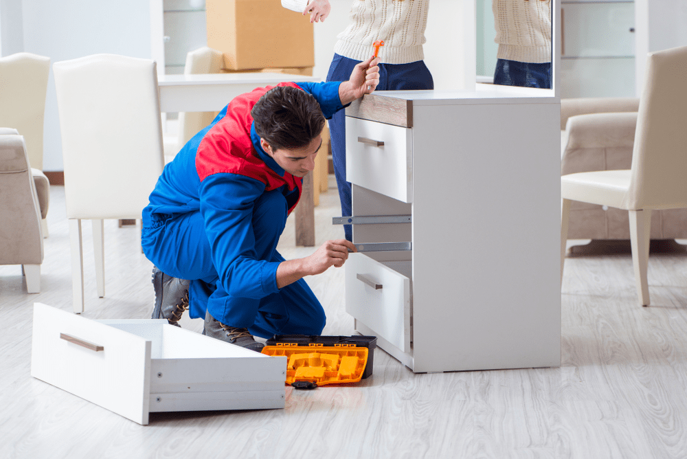 A man in a blue and red jacket kneeling on the floor, fixing a drawer in a white cabinet with a screwdriver.
