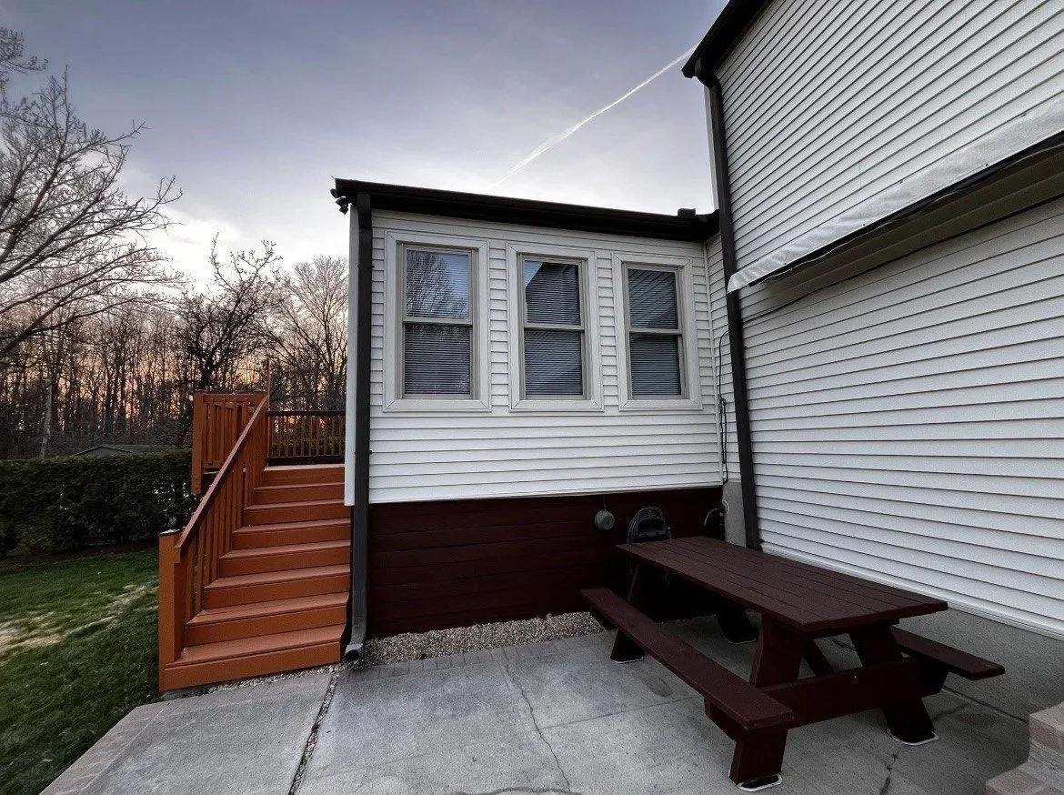 Backyard patio area with a brown picnic table, a small set of red wooden stairs leading to a deck, and white siding on the house at sunset with trees in the background.