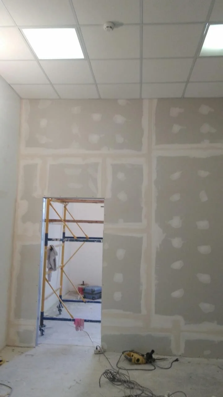 Interior room under construction or renovation with drywall walls and ceiling tiles, construction scaffolding in doorway, and tools on the floor.