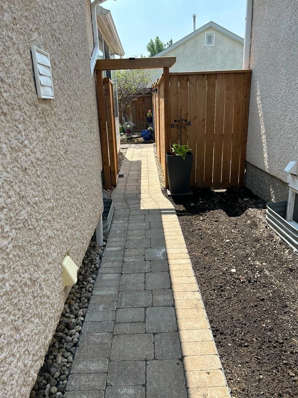 A narrow backyard pathway made of gray brick pavers, bordered by gravel and mulch, leads to a wooden gate with a small tree and potted plants visible beyond.